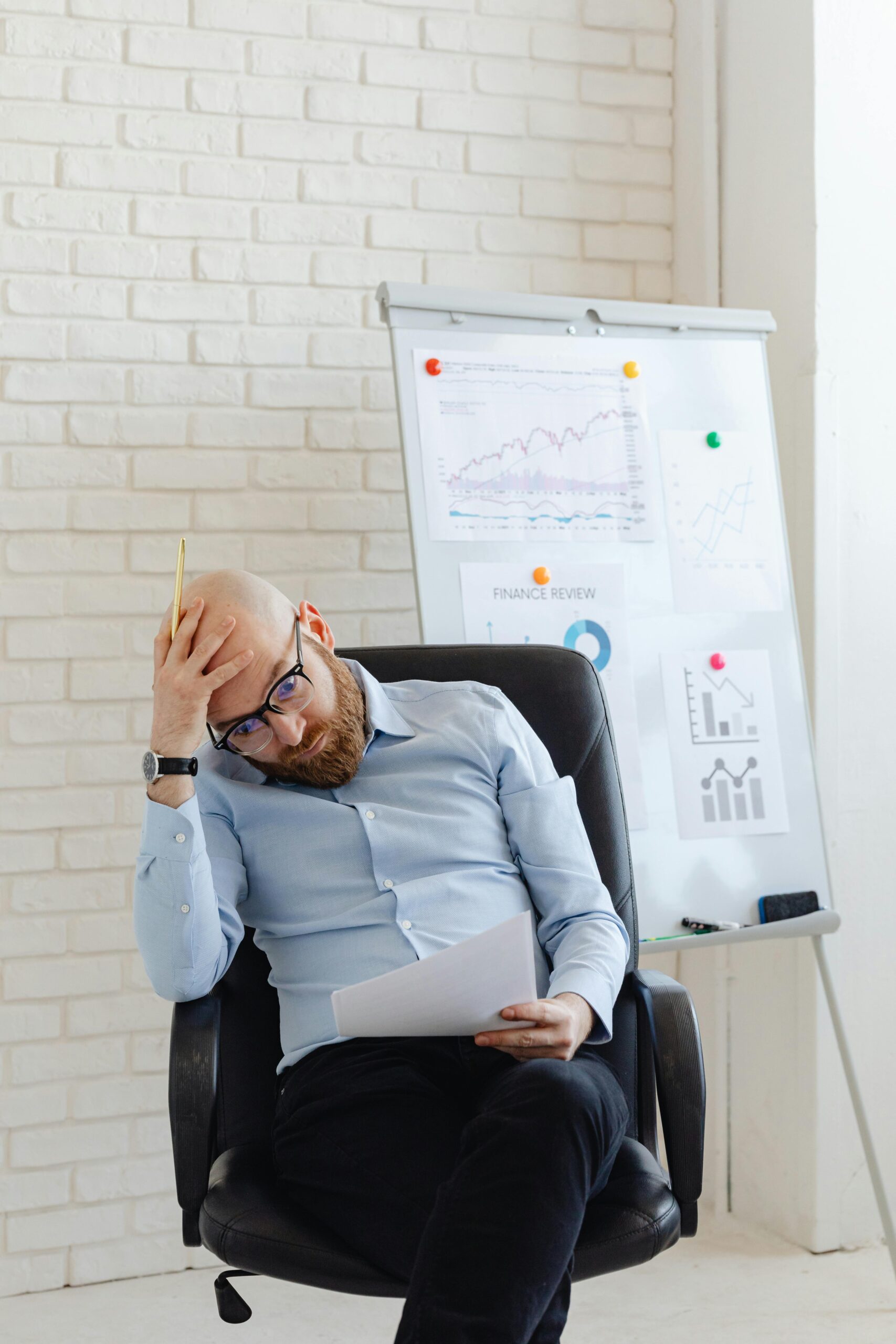 A bald businessman sitting in an office, stressed over financial charts and reports.